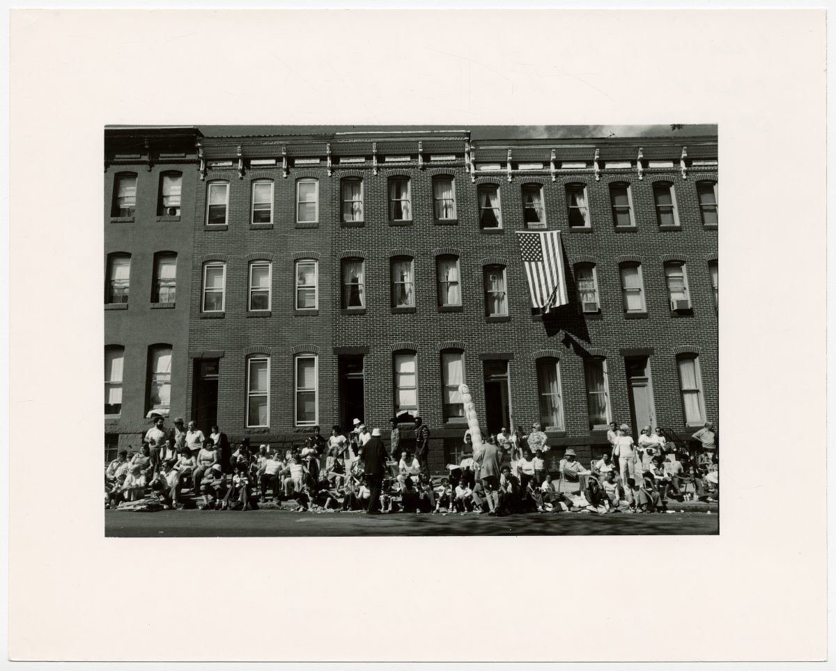 People sitting on stoops and street curb watching the parade. An American flag hangs from a window.