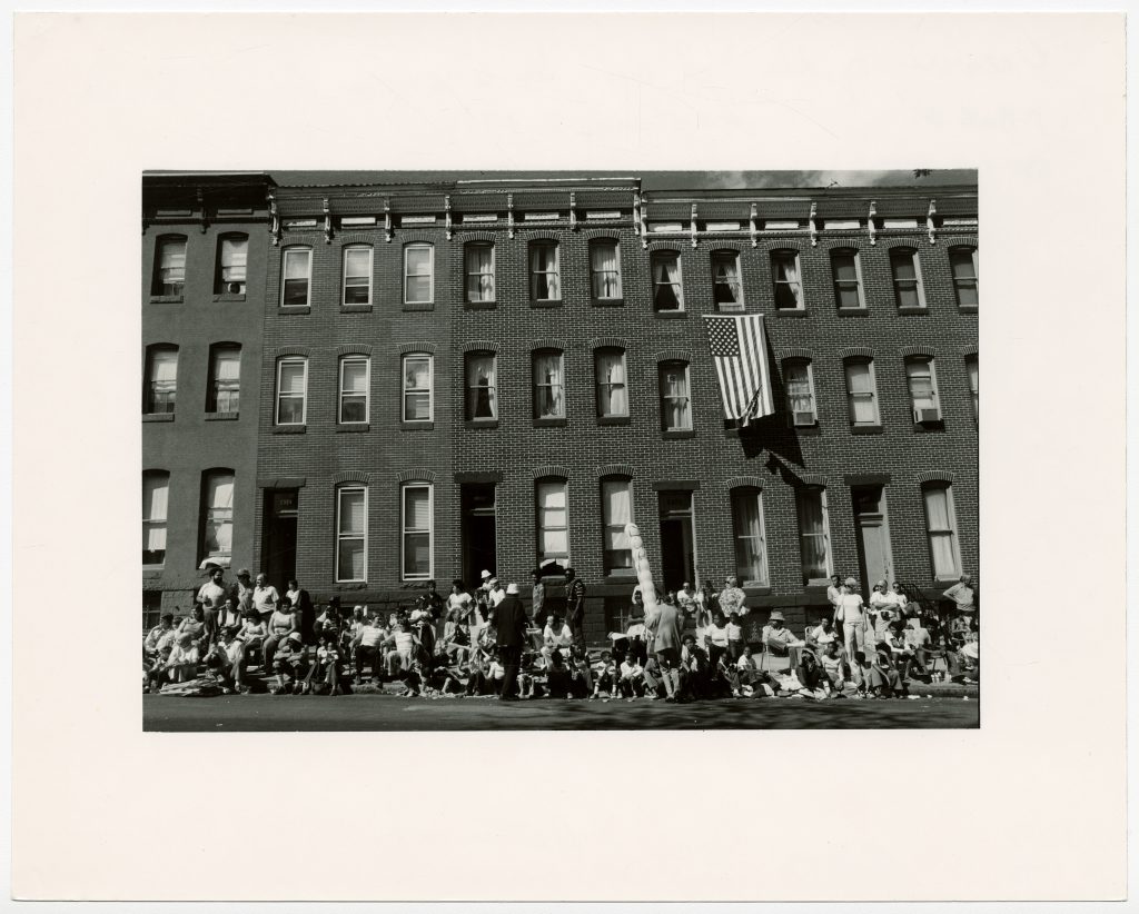 People sitting on stoops and street curb watching the parade. An American flag hangs from a window.