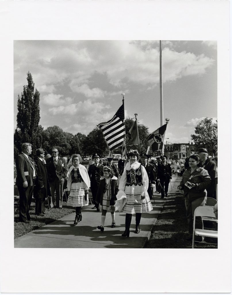 Children walking in a parade with flags behind them and crowds on either side