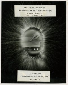 An ovaloid Kirlian projection with a finger touching a rectangular device superimposed inside of the projection. The title and author information are on papers laying on above and below the image, typed in a monospaced font.