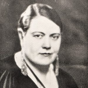 A white woman with short, slicked hair, a black top, and sparkling earrings faces to the right with her head and eyes facing the camera.