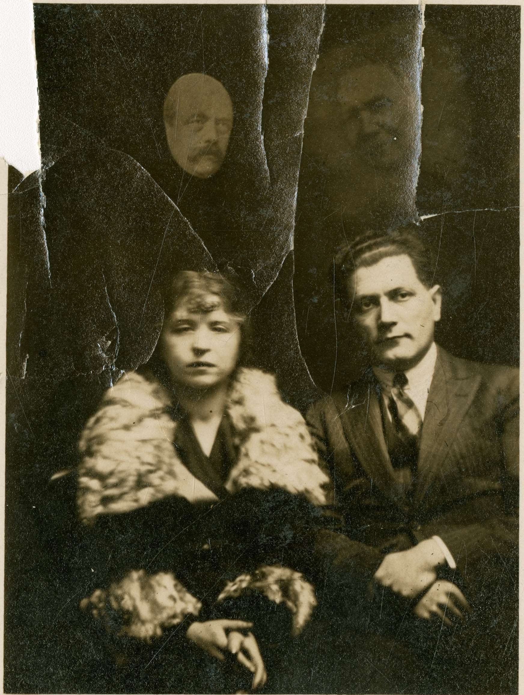 Formal portrait photograph of Nandor Fodor and his wife. Two faces of older people appear in the dark background behind them. 