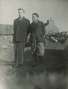 A black and white photograph of two boys wearing warm clothes and standing outside. The boy on the left appears more serious for the photograph, and the boy on the right is looking at him with a joking smile.