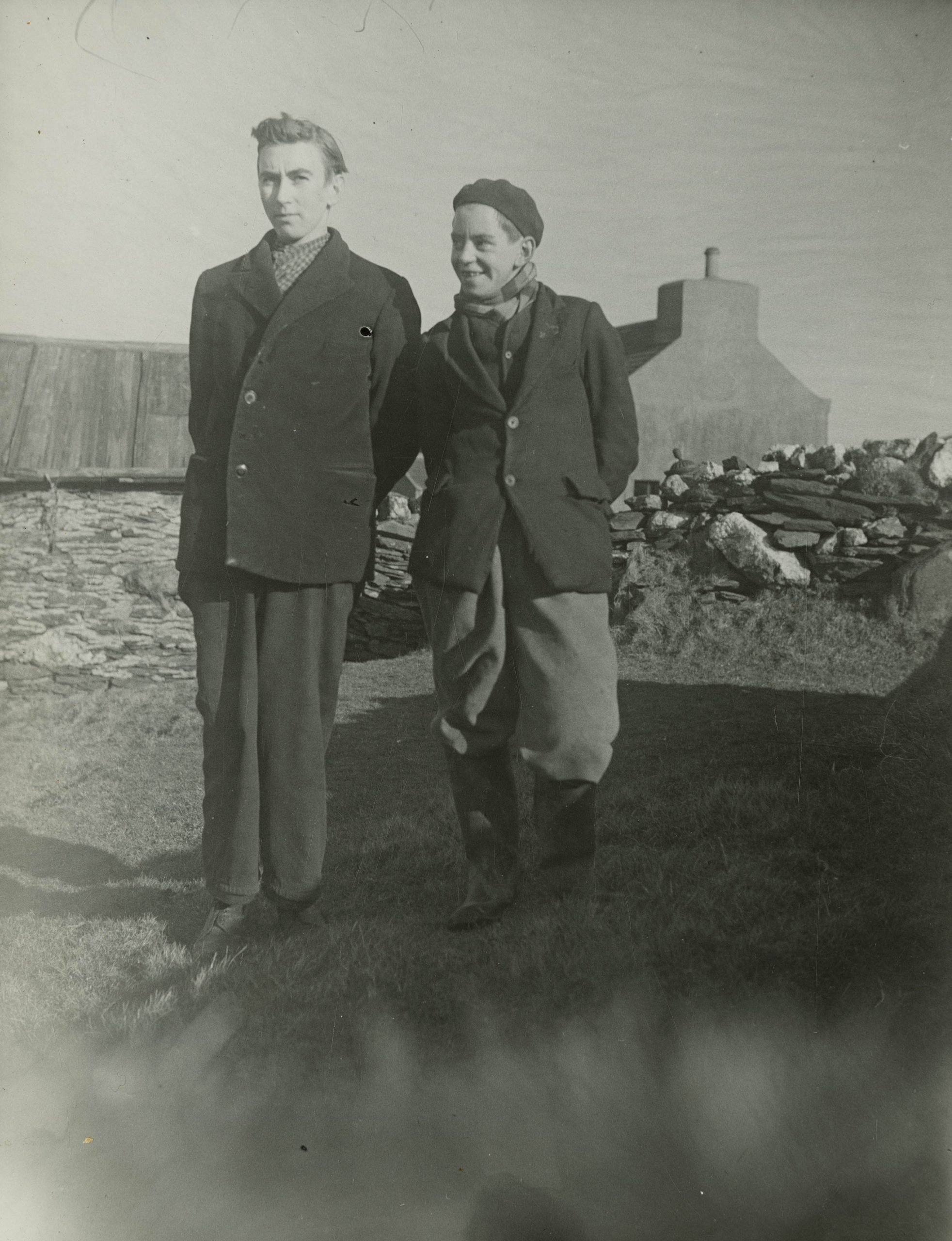 A black and white photograph of two boys wearing warm clothes and standing outside. The boy on the left appears more serious for the photograph, and the boy on the right is looking at him with a joking smile.