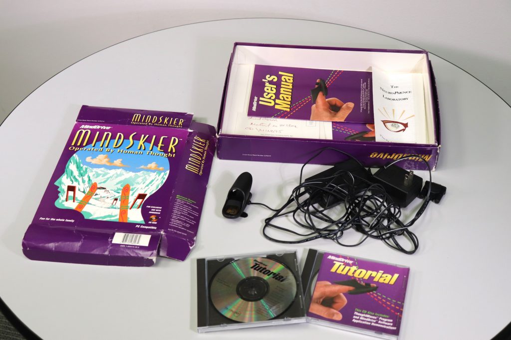 Top: Open box full of paper material. Left: Box that had been flattened with a printed image of a first-person view of skiing cut imposed on the shape of a head. Right: Tangled wires attached to a finger attachment, an HDMI port, and a two-pronged electrical cord. Bottom: two CD cases, on showing the disk inside while the other has a printed paper of a finger on its front.