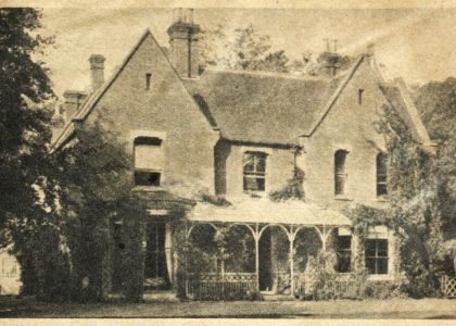 A Gothic-style house with two floors, three chimneys, and a front porch. The house is flanked by trees and slightly overgrown.