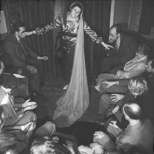 A woman stands with her eyes closed, surrounded by a group of seated men and women. The woman's arms are outstretched and a long piece of fabric is draped from her head, touching the floor.