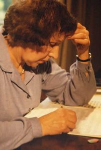 An older white woman with short, curly hair writing with one hand while the other is on her head