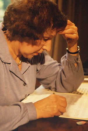 Older woman with short, curly hair hunched over a table as she writes  with one hand. Her other hand is held against her forehead.