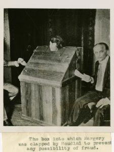 A white woman sits inside a wooden box with multiple locks. Each side of the box has a hole for her hand to poke through. On either side of the box sits a person holding the woman's hand. The person on the left is mostly cropped out of the image. The person on the right is an older white man in a dark suit and tie with short slicked hair. The caption below the image reads: "The box into which Margery was clapped by Houdini to prevent any possibility of fraud."