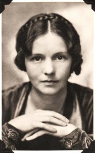 A white woman with short, dark hair and a satin top looks into the camera with her hands clasped beneath her face