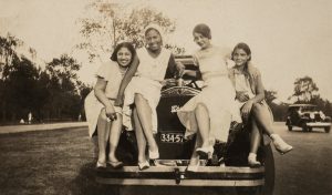 four women sitting on the front of a car.