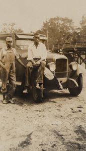 two men in front of car, one sitting on the front fender.