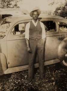 Man in vest and hat standing in front of car looking at camera.