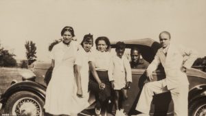 group of adults and child standing in front of car, man sitting in driver's seat.