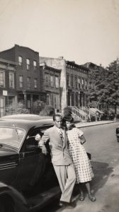 Man and woman looking off-camera leaning against car on neighborhood street.