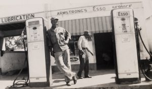 two men in front of Esso gas station, man in front is leaning against gas pump.