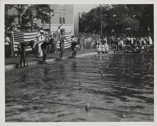 Swim meet featuring four youth diving into a pool, surrounded by onlookers outside a fence rimming the pool.