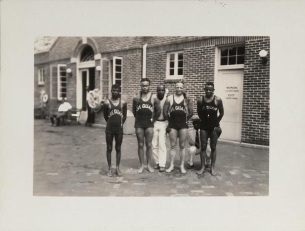 Four young people stand shoulder to shoulder, looking into the camera. They are wearing swimsuits printed with the word "lifeguard" across the chest. Behind them, one man stands and one map crouches. 