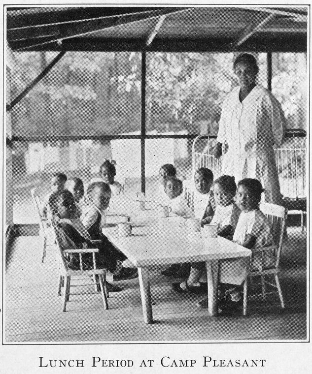 A group of young Black children sit around a table, with an adult woman counselor standing on the right.