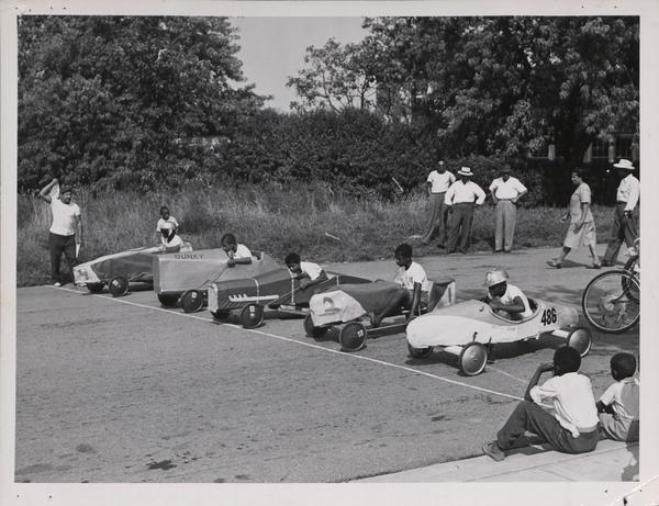 The starting line of a soapbox derby race with five children in cars, surrounded by seven onlookers