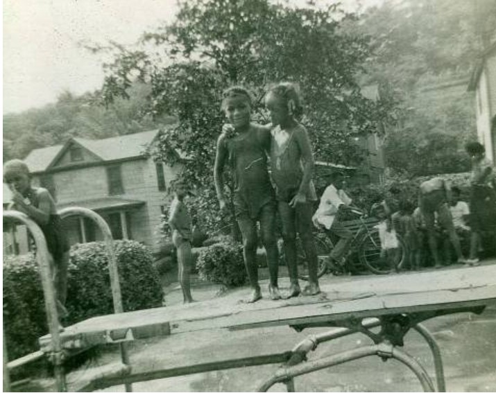 Two girls pose on diving board. Several people gather in the background, including one man on a bicycle.