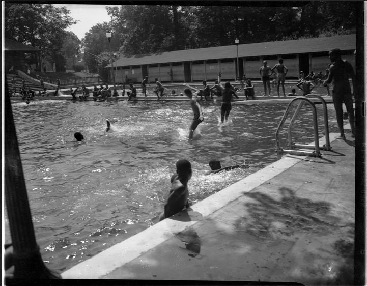 Summer scene at Druid Hill Pool Number 2 with people swimming, jumping into the water, and standing around the pool.