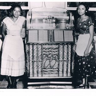 Two women stand on either side of jukebox.