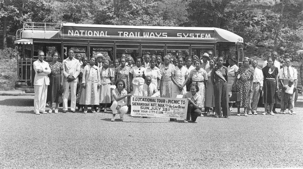 A group of people stand in from of a bus. They are holding a sign reading "Educations Tour, Picnic To Shenandoah Natl Park via Skyline Drive. Sun. July 25th. Know Your City Club"