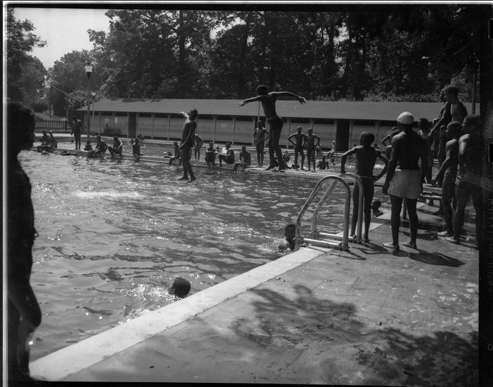 Scene at Druid Hill Pool Number 2 featuring two people jumping off a diving board, several people swimming, and others standing around the pool.