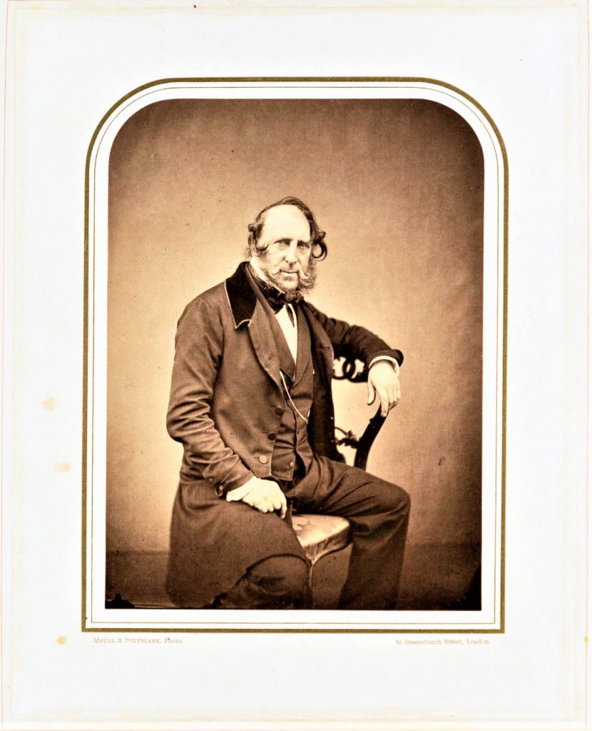 A white man with curly hair sits in a chair. he is wearing a dark suit and has his arm on the back of the chair. 